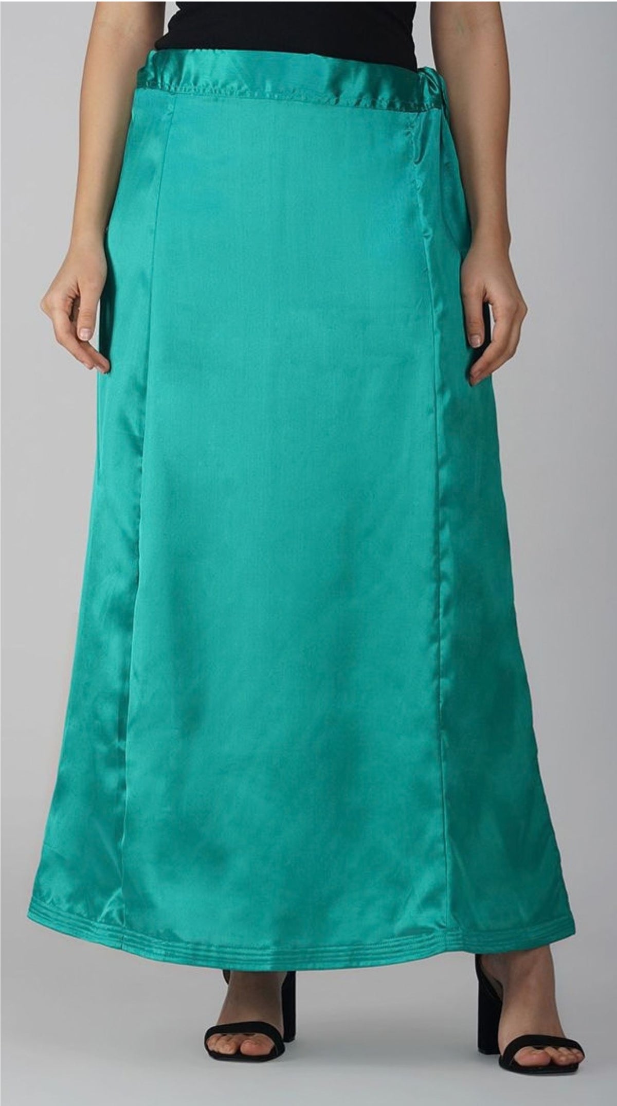A model wearing a long, flowing teal satin skirt with side pockets, paired with black heeled sandals against a neutral gray background.