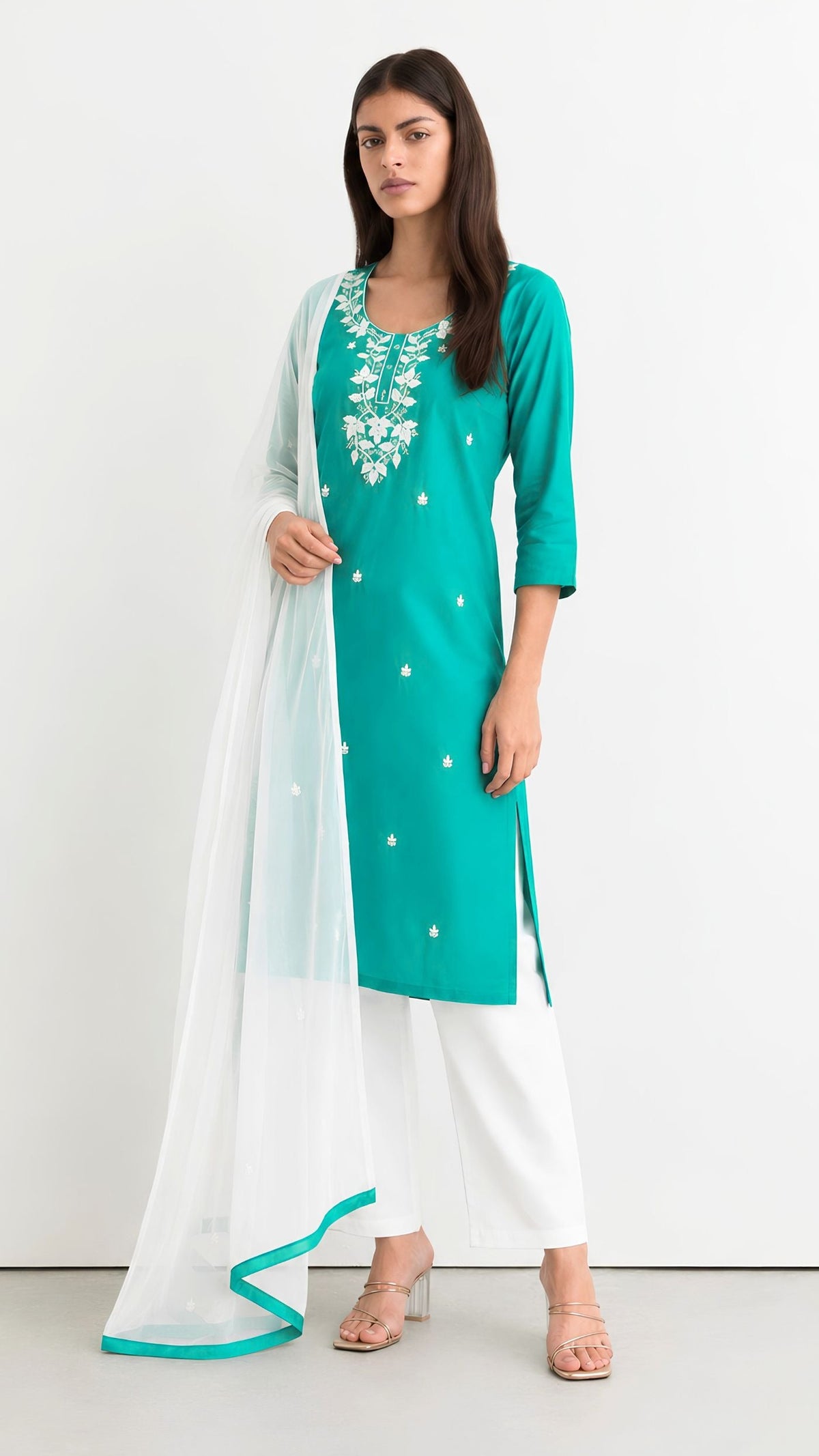A woman modeling a teal green kurta with white floral embroidery, paired with white pants and a sheer white dupatta with a teal border, set against a plain white studio background.