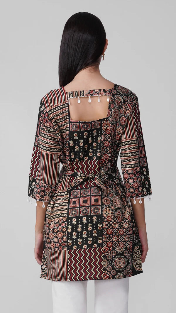 A woman shown from the back wearing a patterned tunic top with three-quarter sleeves, a square neckline with tassel details, and a tie waist, paired with white pants against a light gray studio background.