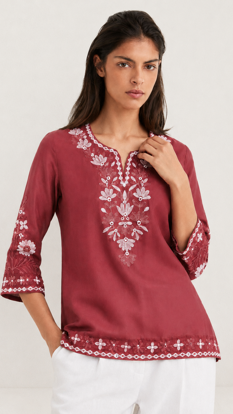 A woman modeling a three-quarter sleeve maroon tunic top with white floral embroidery on the front, neckline, and cuffs, paired with white pants against a plain light background.