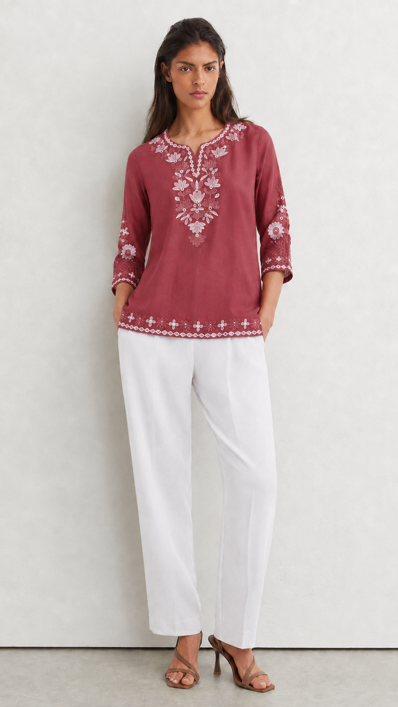 A woman modeling a dusty rose embroidered tunic top with white wide-leg pants and brown heeled sandals, standing against a light gray textured wall.