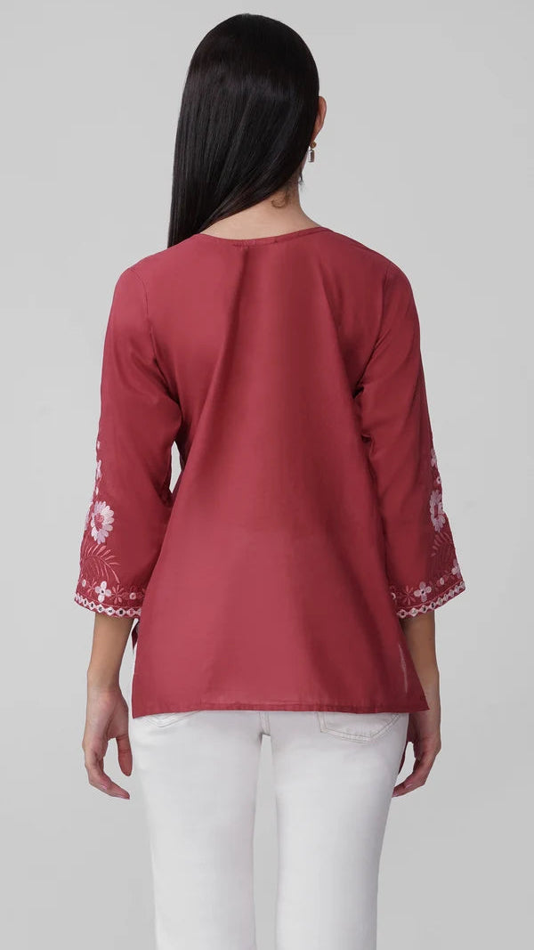 A woman viewed from the back wearing a dusty rose red three-quarter sleeve top with white floral embroidery on the cuffs, paired with white pants, against a light gray studio background.