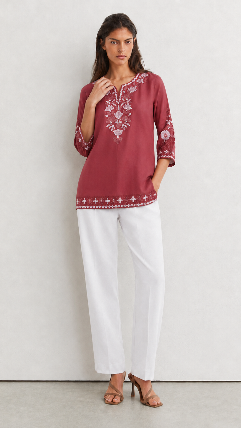 A woman modeling a dusty rose embroidered tunic top with white wide-leg pants and brown heeled sandals, standing against a light gray wall.