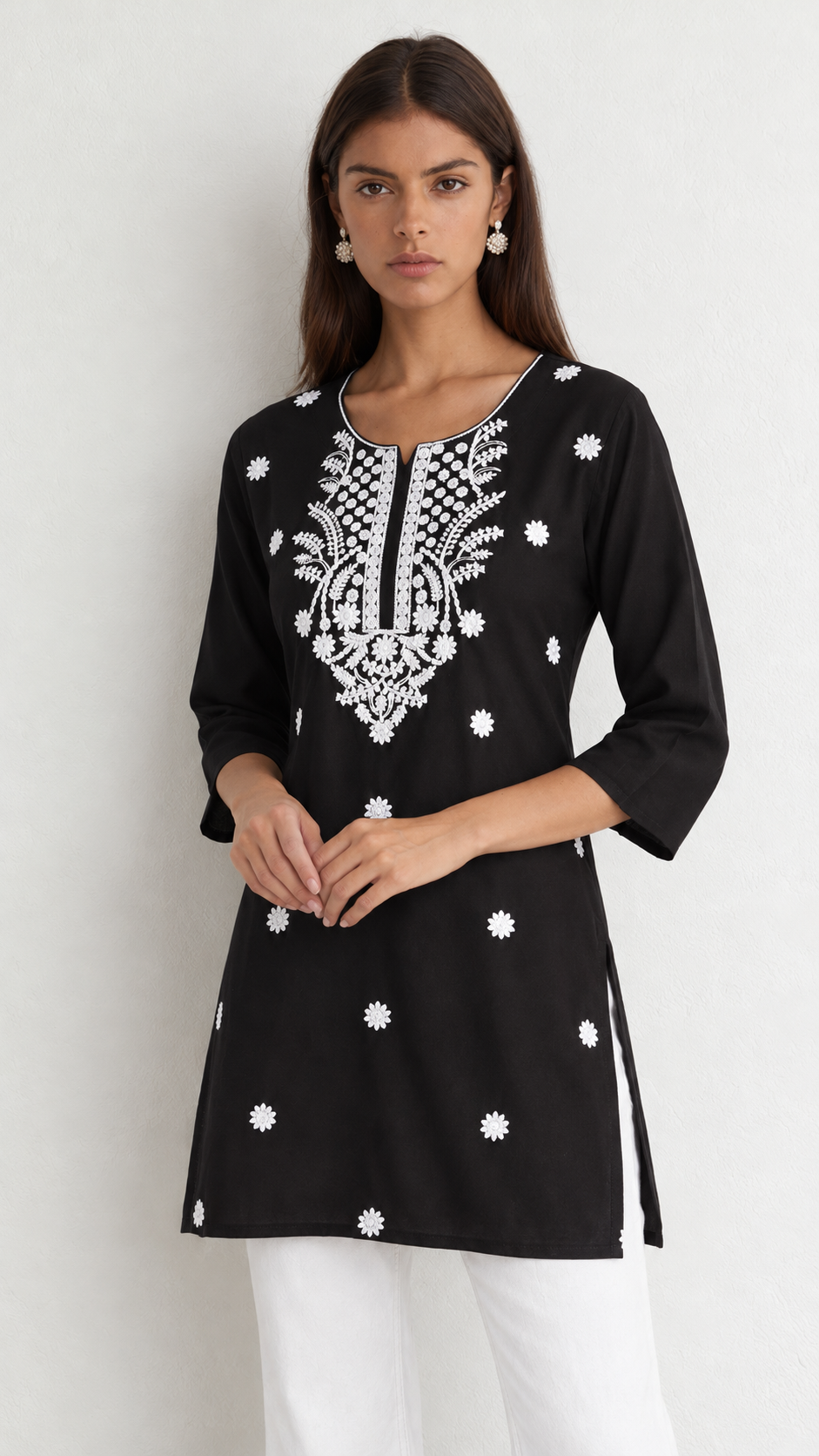 A woman modeling a black three-quarter sleeve kurta with intricate white embroidery on the chest and scattered white floral motifs, paired with white pants against a plain light gray background.