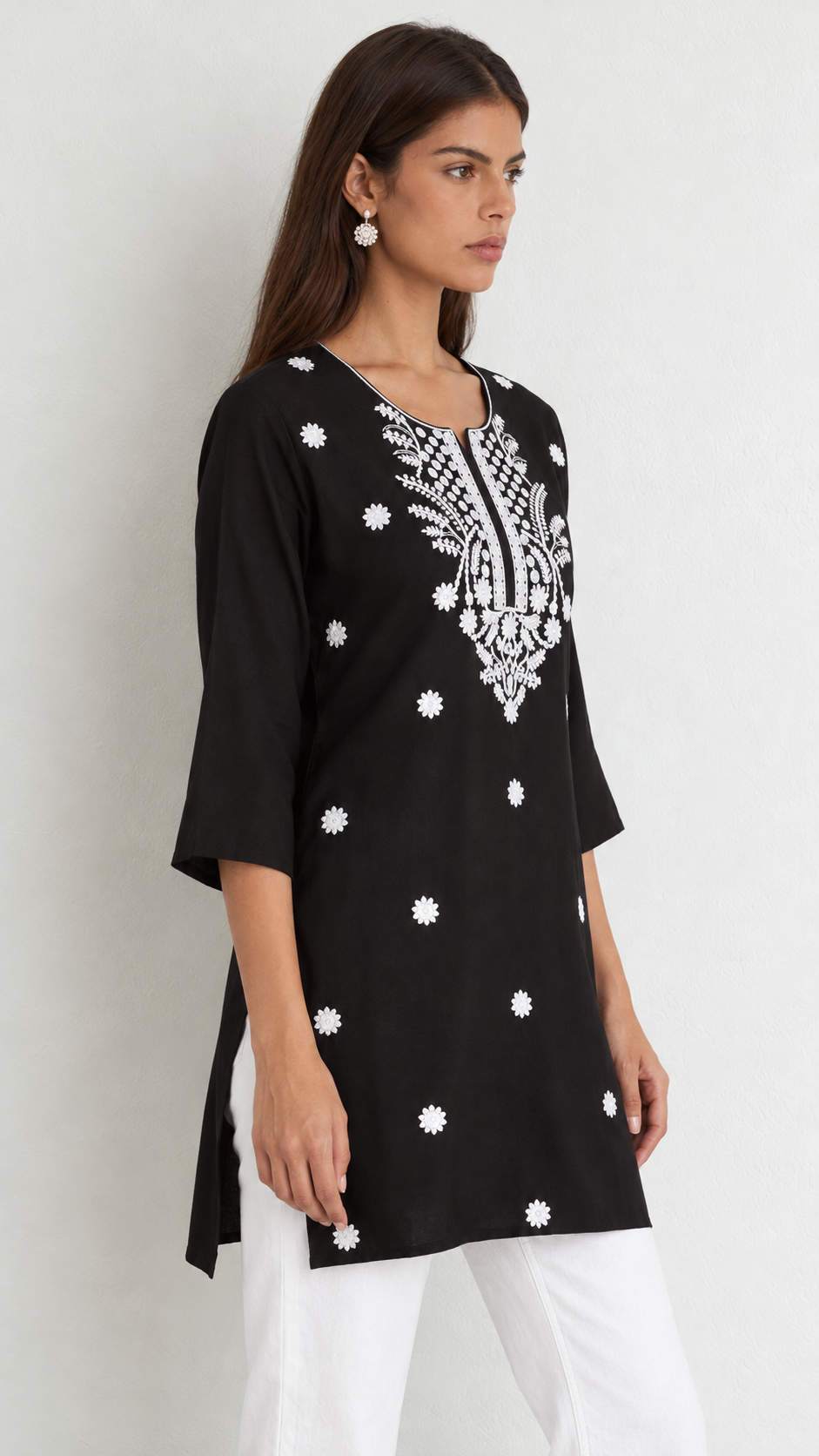 A woman wearing a black three-quarter sleeve kurta with white floral embroidery and white pants, standing against a light gray studio background.