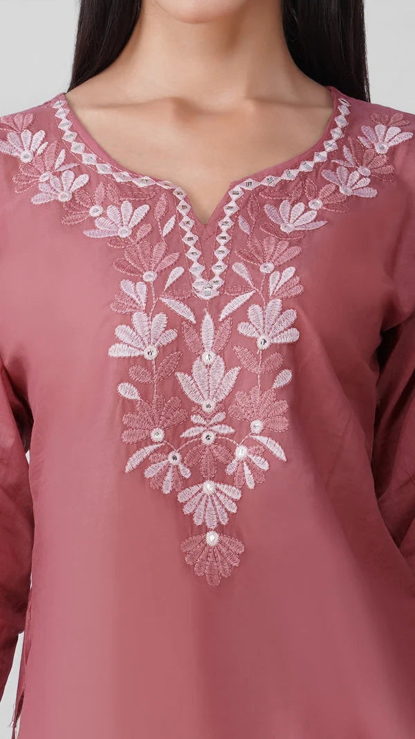 Young woman wearing a dusty rose embroidered kurta top with white floral threadwork, three-quarter sleeves and white flared trousers.