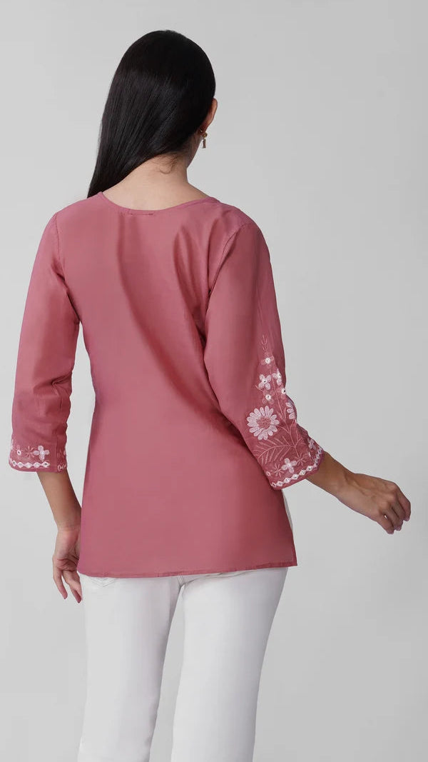 Young woman wearing a dusty rose embroidered kurta top with white floral threadwork, three-quarter sleeves and white flared trousers.