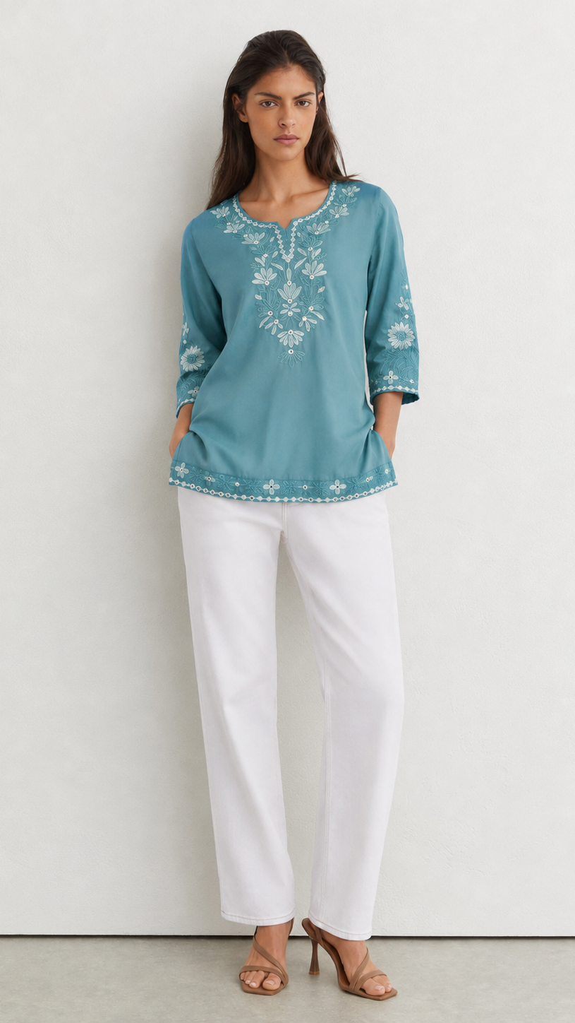 A woman modeling a teal blue embroidered tunic top with white pants and tan heeled sandals, standing against a plain off-white wall.