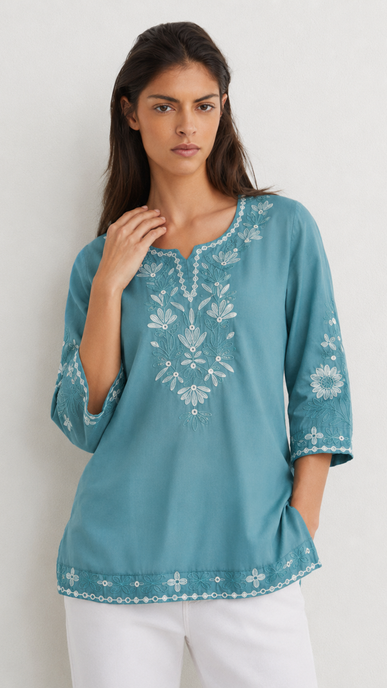 A woman wearing a teal blue embroidered tunic top with white floral patterns and white pants, standing against a plain off-white wall.