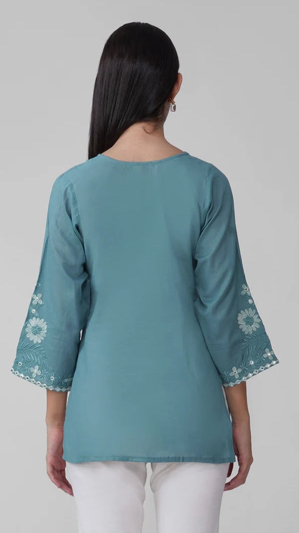 A woman viewed from the back wearing a teal blue-green tunic top with white floral embroidery on the three-quarter sleeves, paired with white pants, against a neutral light gray background.
