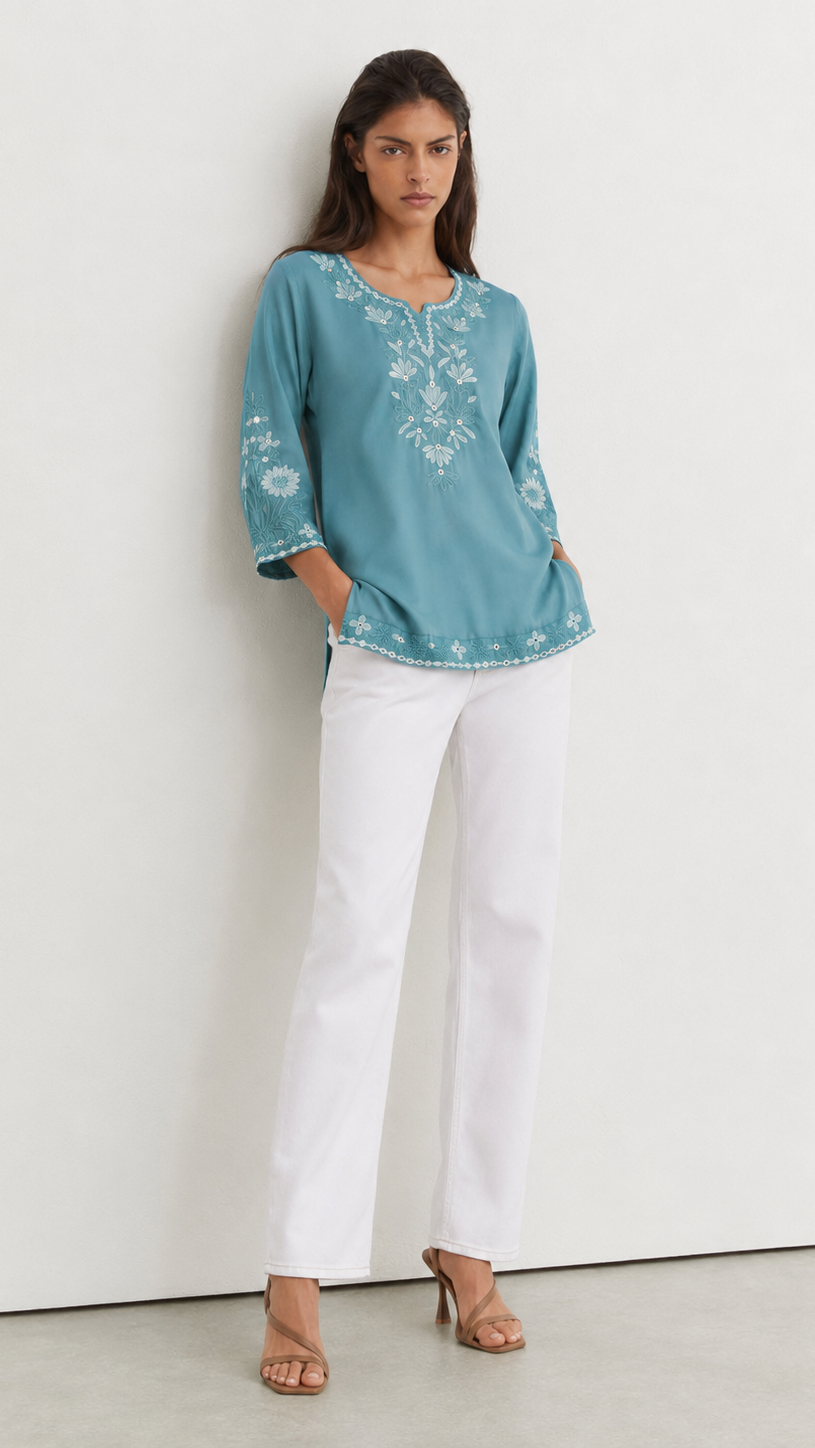 A woman stands against a plain white wall wearing a teal embroidered tunic top with white three-quarter sleeves, white pants, and tan heeled sandals.