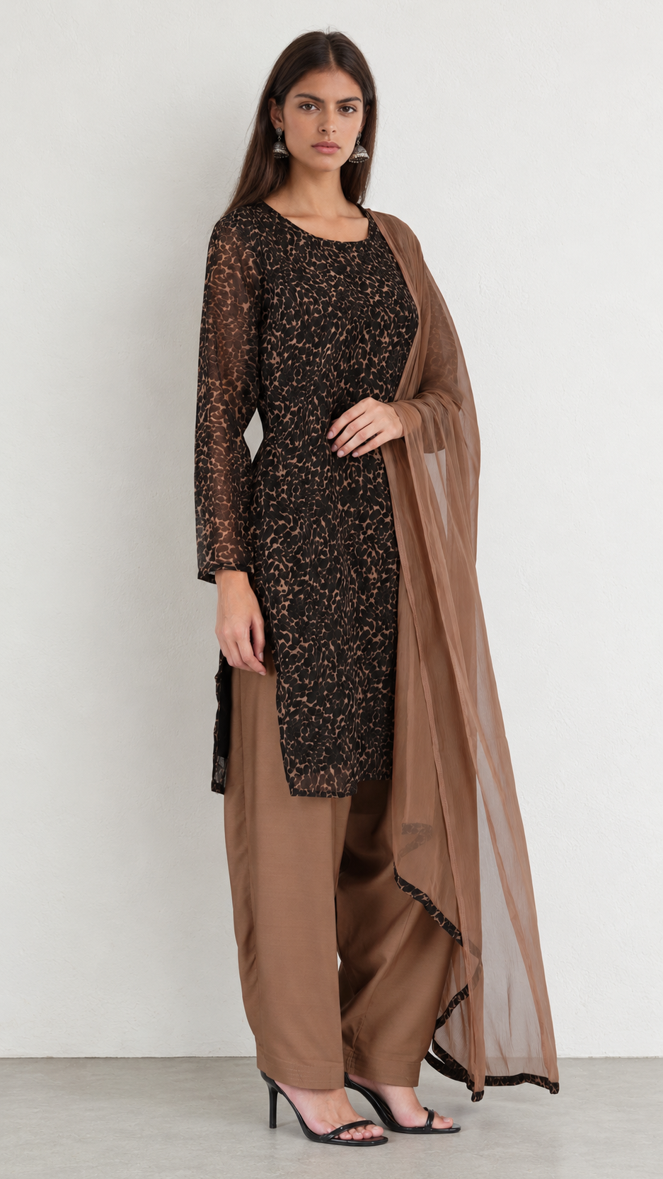 A woman modeling a traditional three-piece salwar kameez set in brown. The outfit features a long-sleeved black and brown patterned kameez top, matching brown trousers, and a sheer brown dupatta, set against a plain light gray studio background.