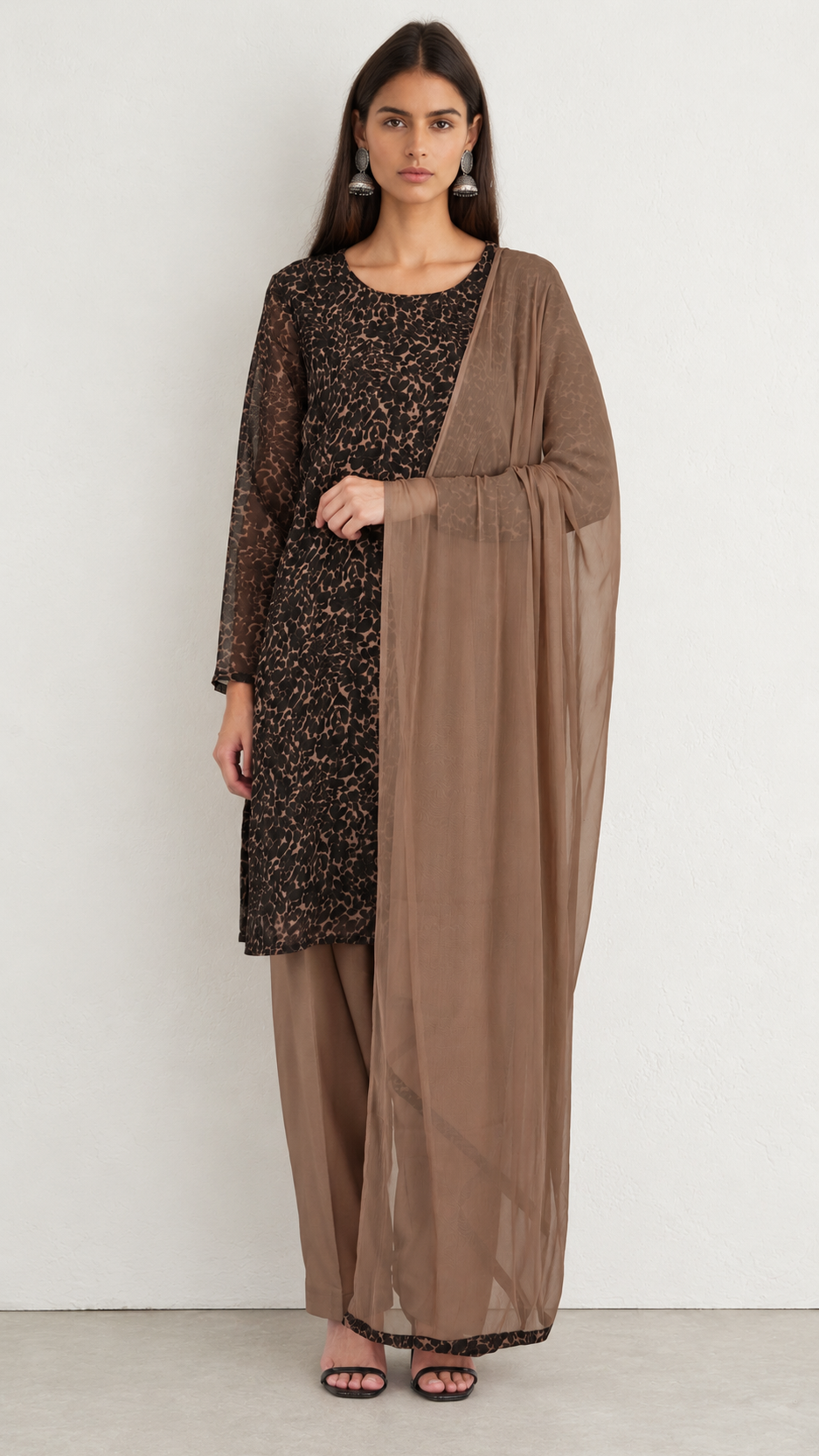 A woman modeling a traditional outfit consisting of a black floral printed kurta, matching wide-leg pants, and a sheer taupe-brown dupatta, set against a plain light gray studio background.