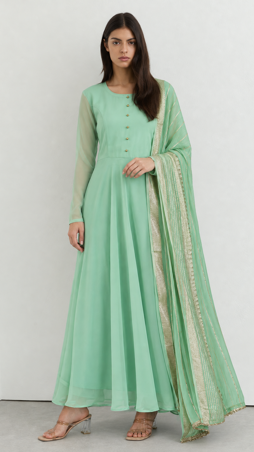 A woman modeling a mint green traditional Indian outfit consisting of a long-sleeved kurta with button details, a matching flared skirt, and a sheer dupatta with gold border detailing, set against a plain white studio background.