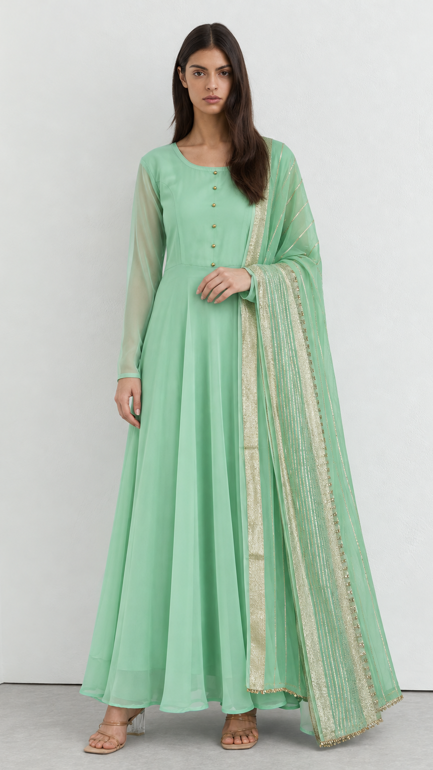 A woman modeling a mint green anarkali-style dress with a matching sheer dupatta featuring gold embroidery, set against a plain light gray studio background.