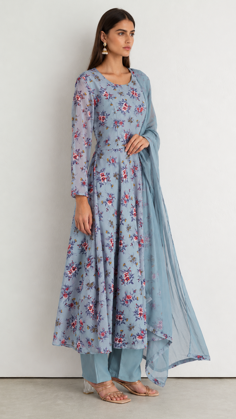 A woman modeling a light blue floral printed kurta set with matching pants and a sheer dupatta, standing against a plain off-white wall.