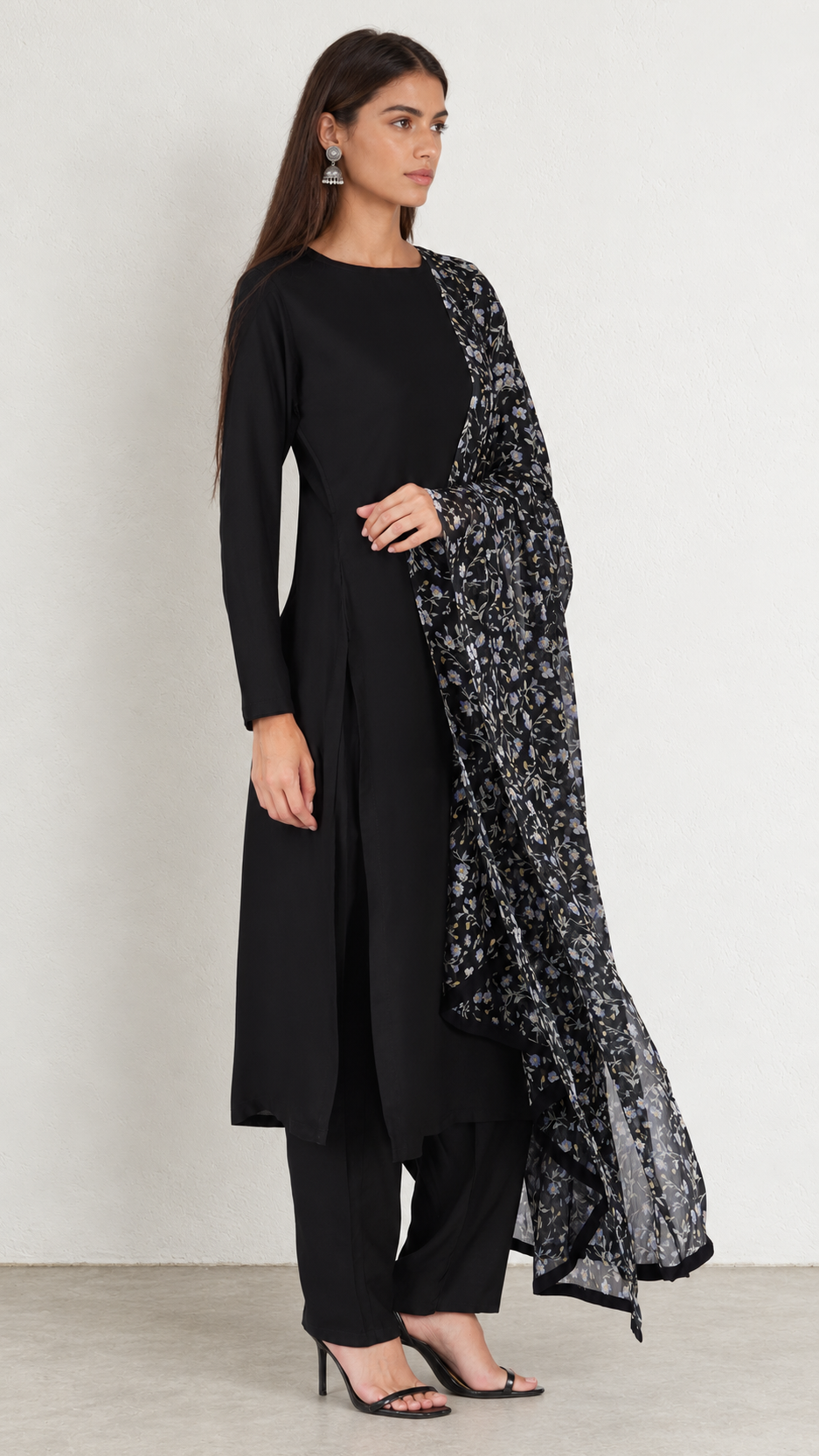 A woman modeling a black long kurta with black pants and a black-and-white patterned dupatta, standing against a plain light gray wall.