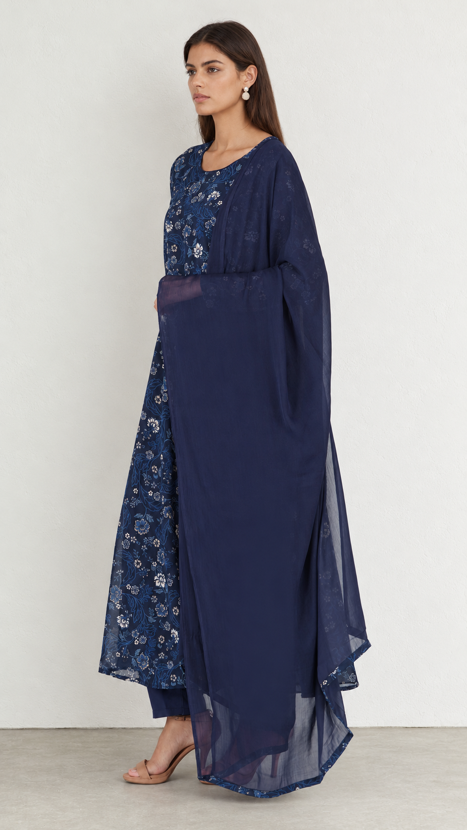A woman modeling a navy blue traditional outfit with a sheer matching outer layer, featuring floral embroidery on the bodice and lower skirt, set against a plain light gray studio background.