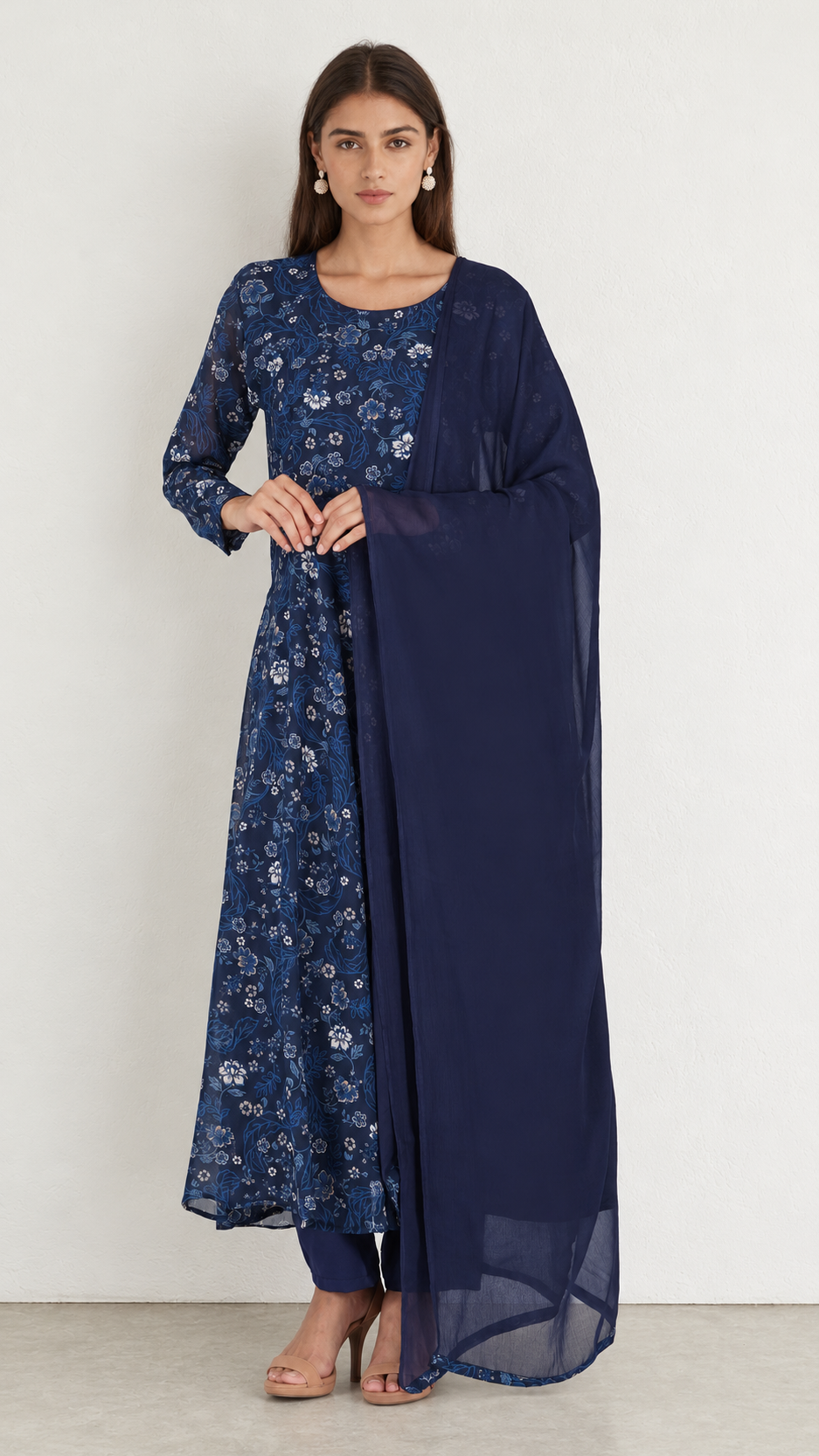 A woman modeling a navy blue traditional outfit consisting of a floral printed kurta, matching trousers, and a solid navy dupatta, standing against a plain off-white wall.