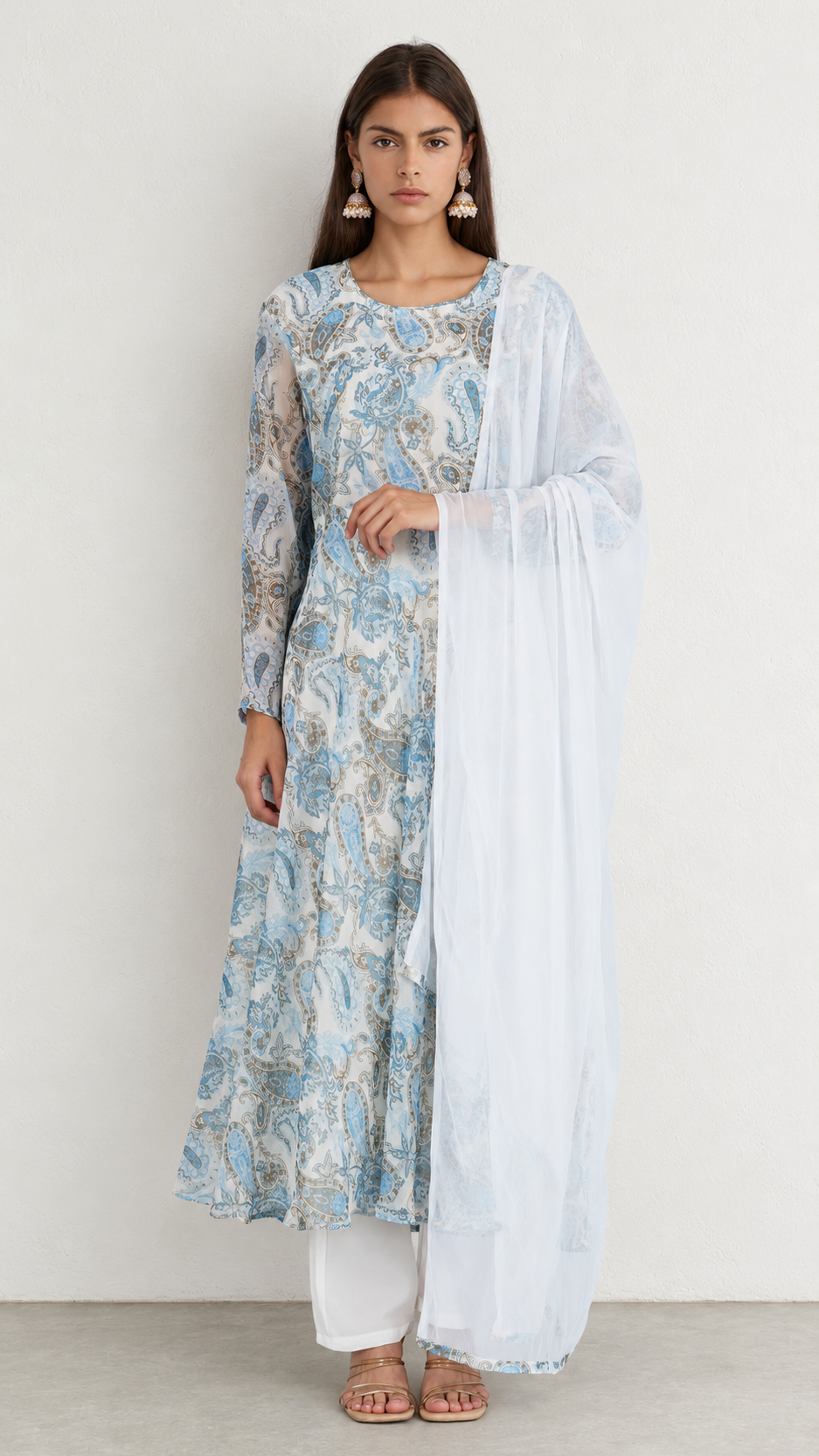 A woman modeling a light blue and white printed kurta set with a white dupatta, white pants, and flat sandals against a plain off-white studio background.