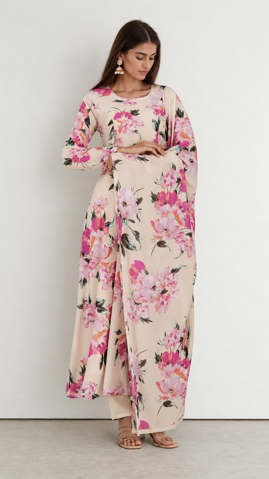 A woman modeling a long-sleeved maxi dress with a pink and green floral print on a beige background, standing against a plain light gray wall.