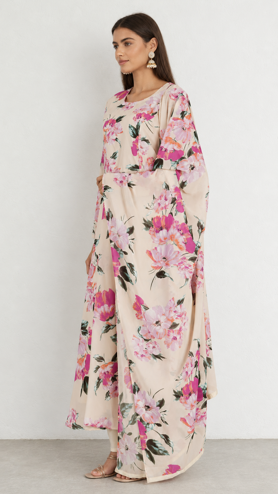 A woman modeling a long, flowing kaftan dress with a pink and green floral print on a beige background, standing against a plain light gray wall.