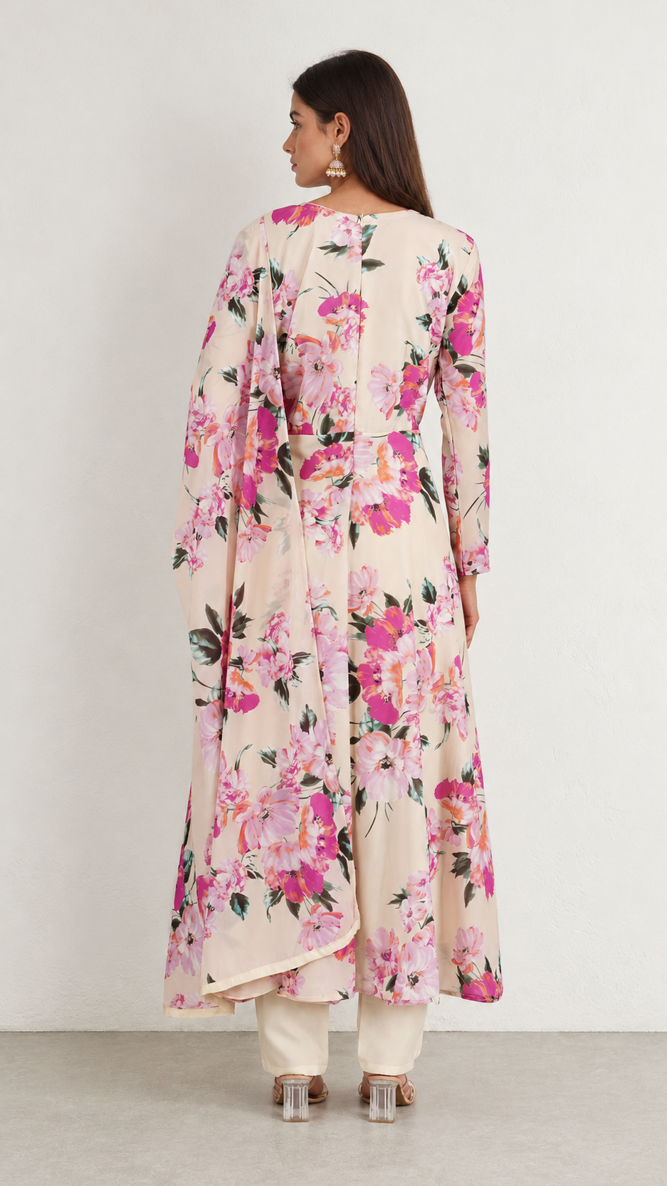 A woman shown from the back wearing a long-sleeved cream-colored kurta with a vibrant pink and green floral print, paired with white pants and clear high-heeled sandals, set against a plain light gray studio background.