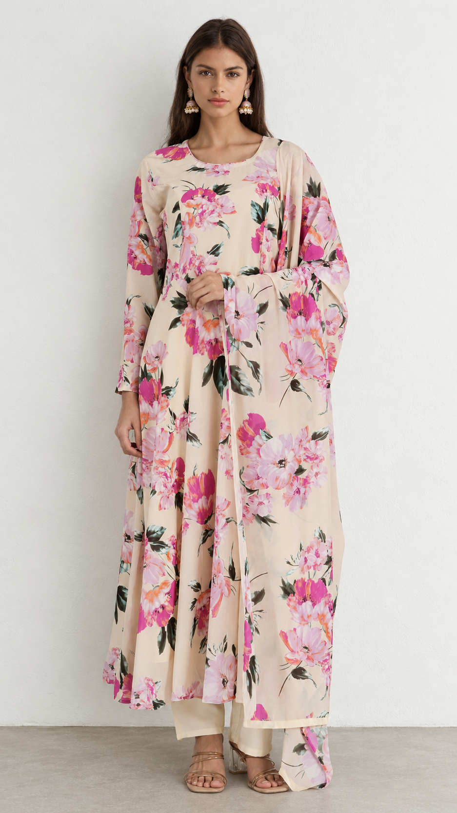 A woman modeling a long-sleeved beige maxi dress with a vibrant pink and green floral print, standing against a plain white wall.