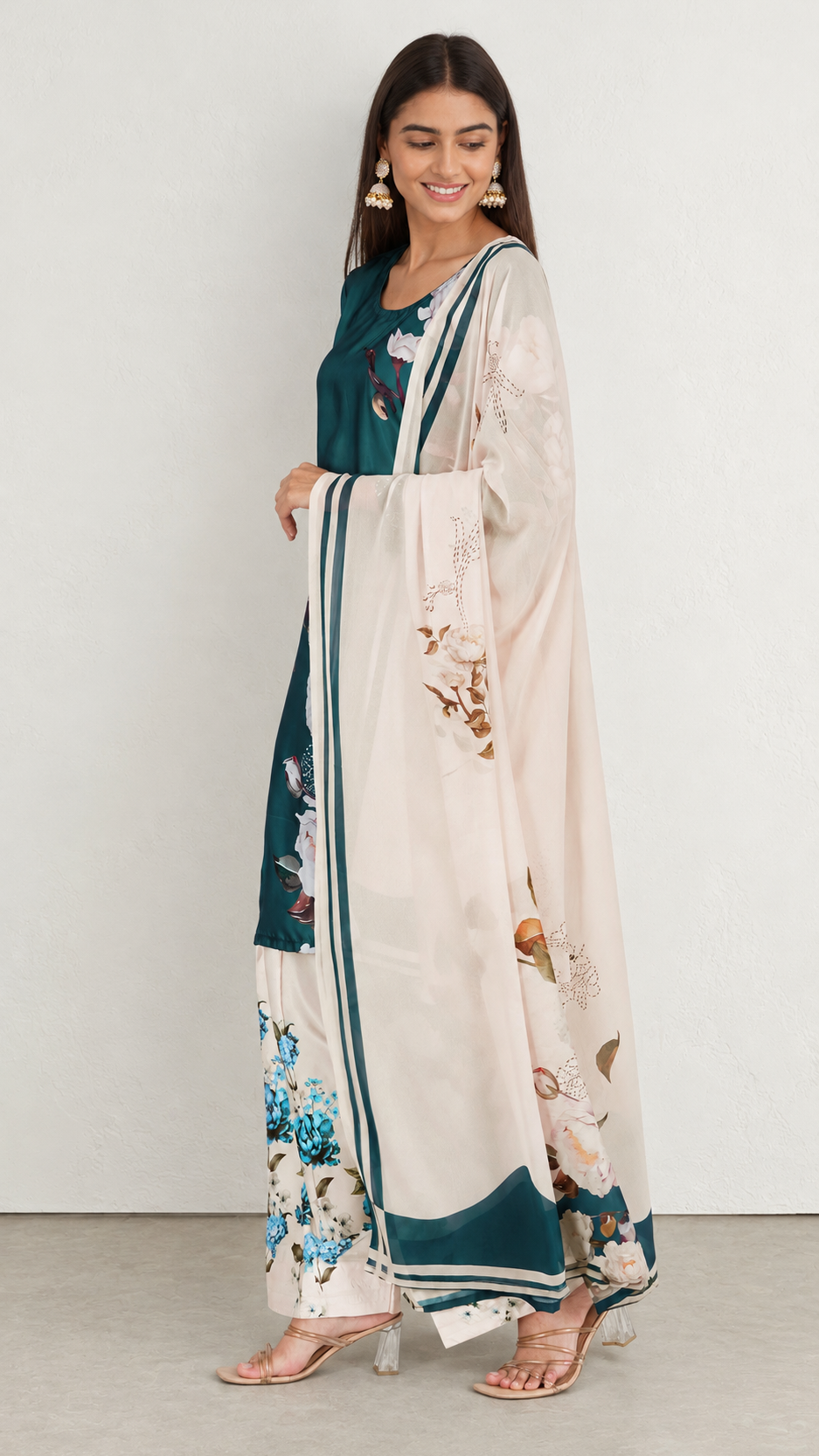 A woman models a traditional three-piece salwar kameez suit. The set features a dark teal green kurta, matching wide-leg trousers, and a large cream-colored dupatta with blue and brown floral prints and green borders.