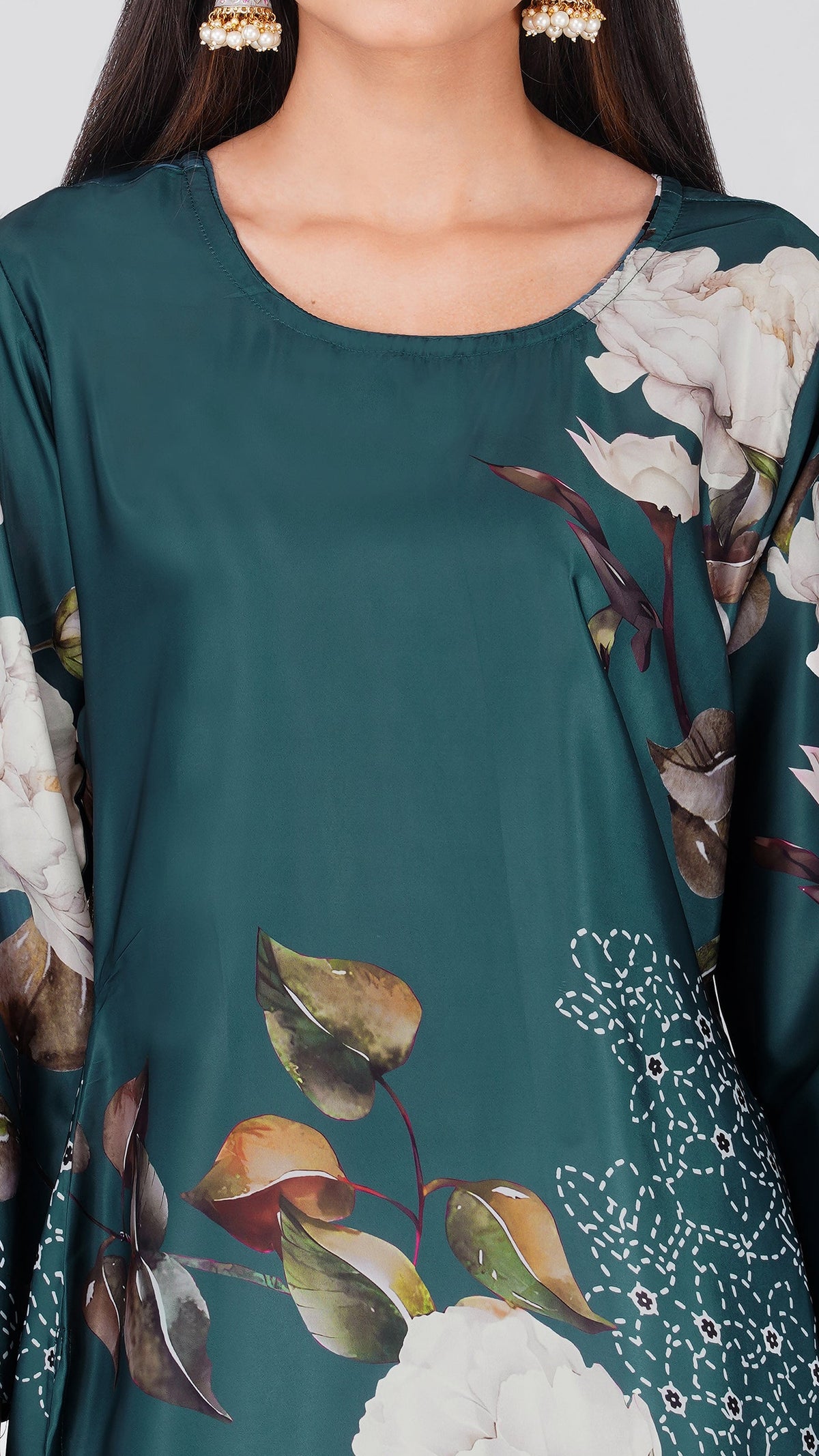 A close-up of a woman wearing a dark teal green tunic top with a large white and brown floral print, styled with gold earrings.