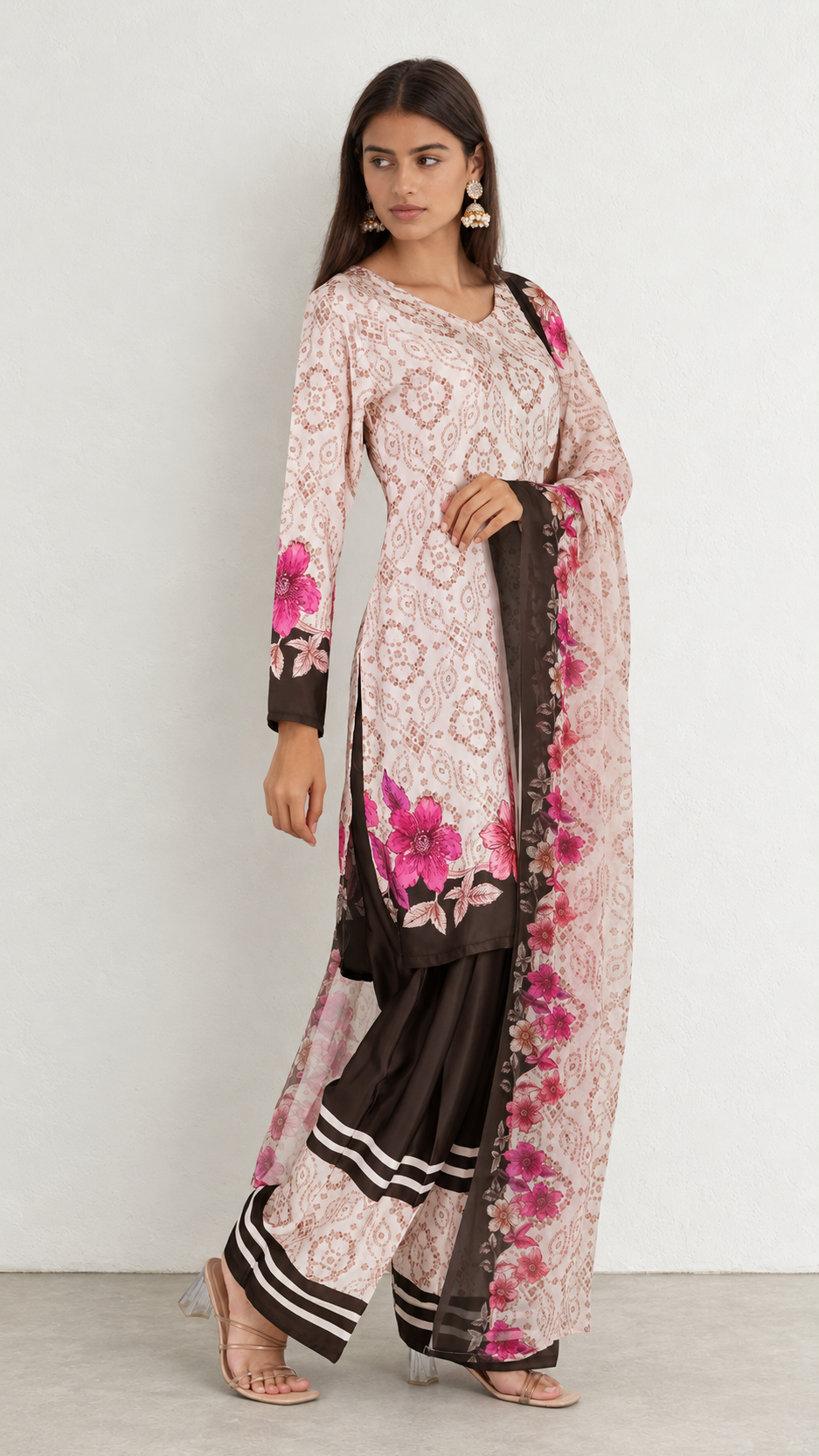A woman modeling a traditional three-piece salwar kameez suit in light pink and brown. The kurta features a pink floral print over a brown base, paired with matching brown sharara pants and a sheer brown dupatta with a pink floral border.