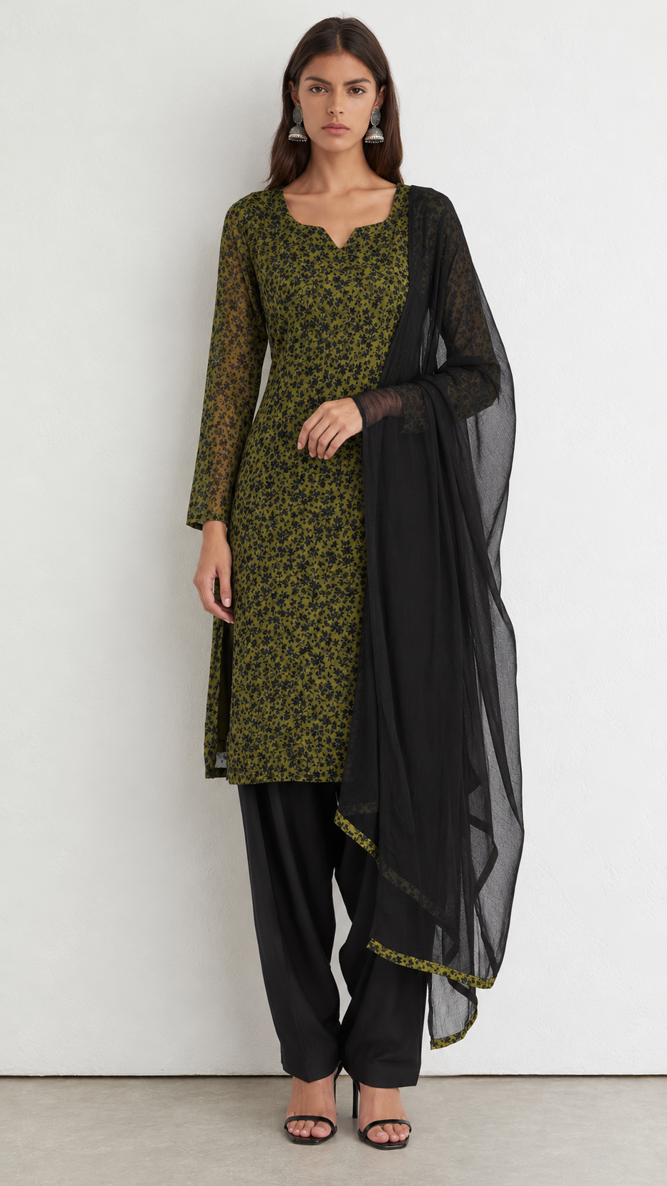 A woman modeling a traditional three-piece salwar kameez set featuring an olive green and black patterned kurta, black trousers, and a sheer black dupatta with a gold border, set against a plain white studio background.