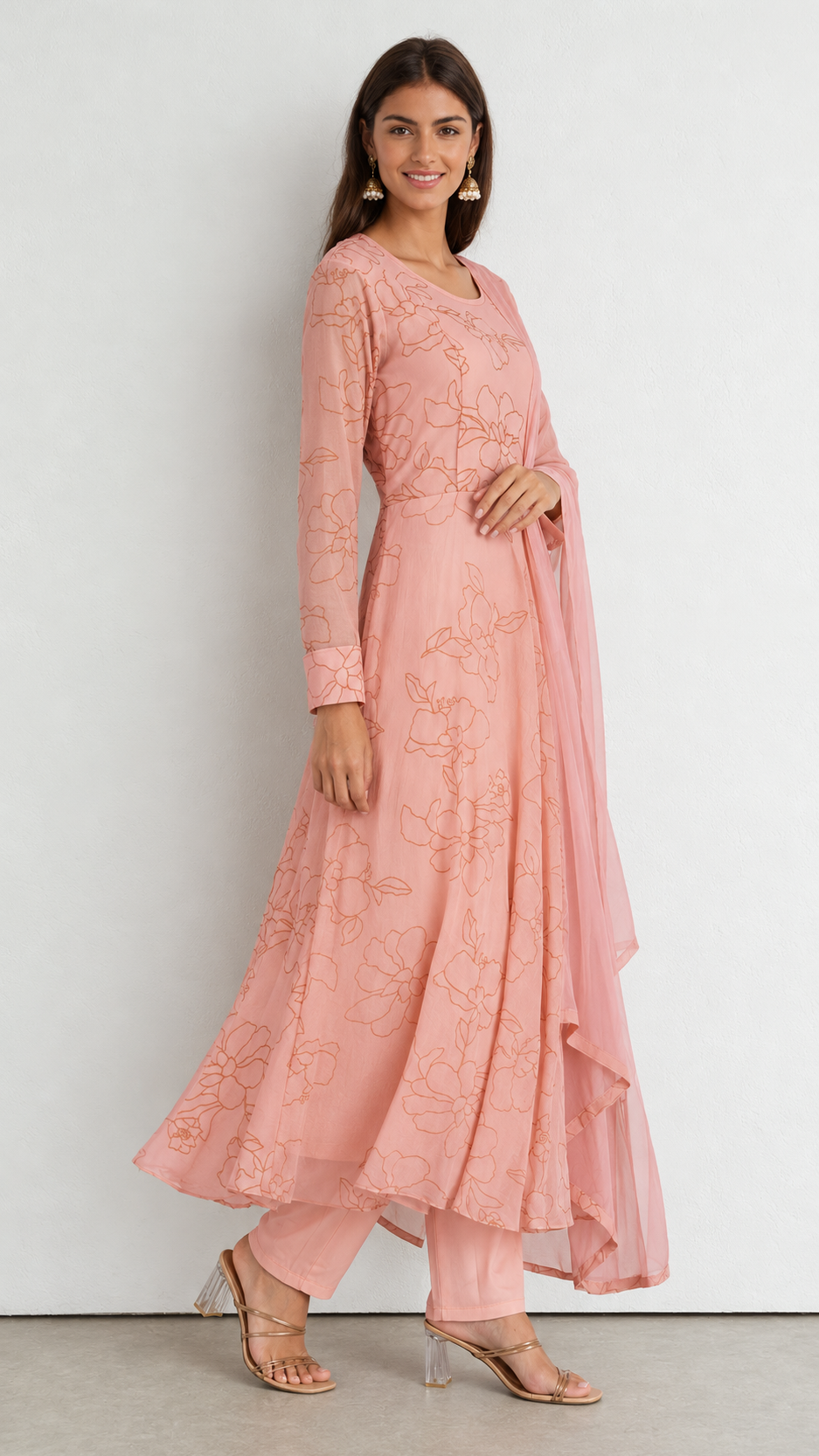 A woman modeling a peach-pink long-sleeve embroidered kurta set with matching pants and a draped dupatta, standing against a plain light gray wall.