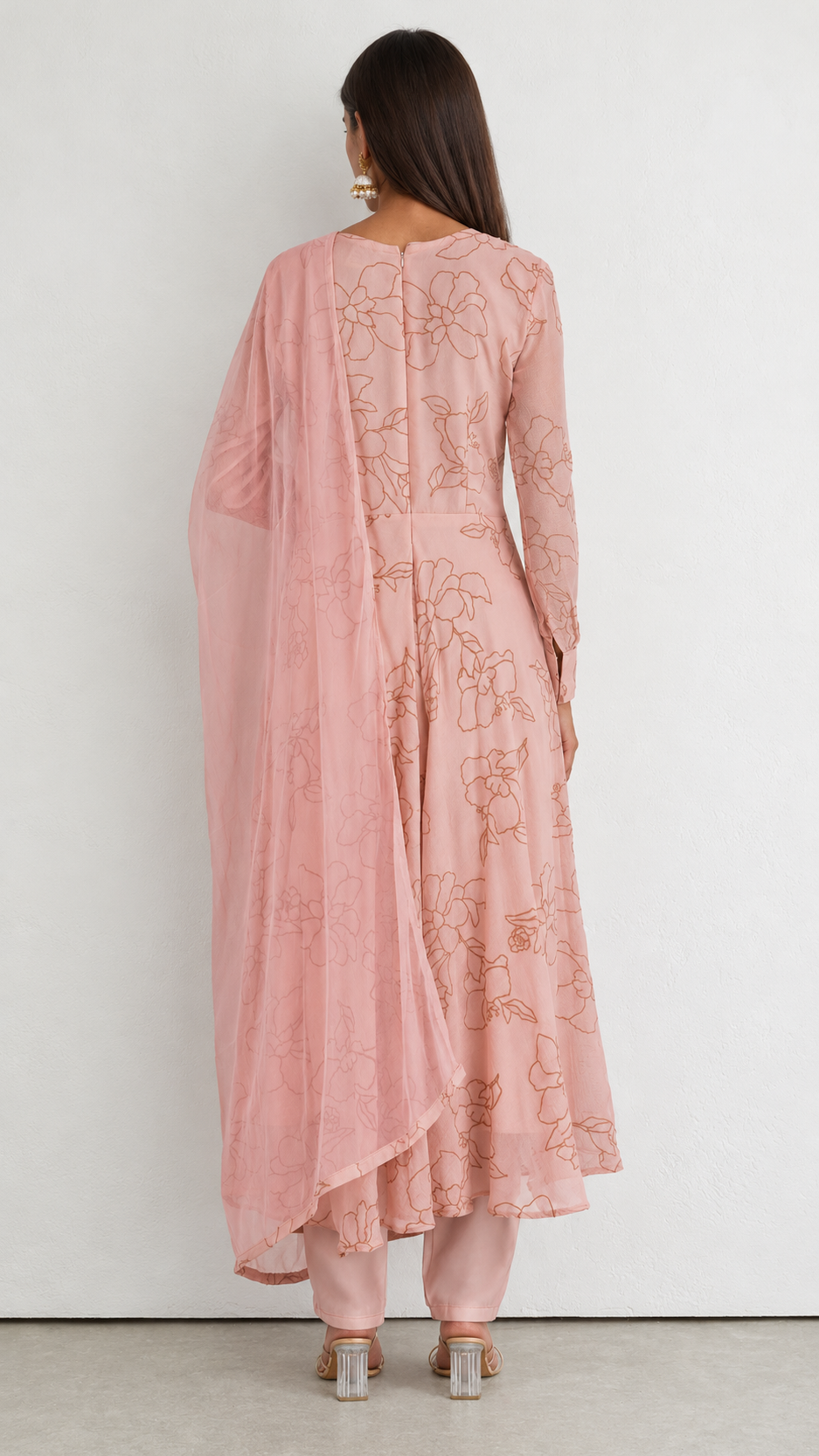 A woman shown from the back wearing a dusty pink traditional outfit with floral embroidery, featuring a long kurta, pants, and a matching sheer dupatta, set against a plain light gray wall.