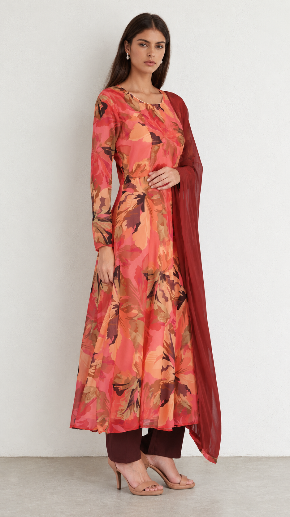 A woman modeling a long-sleeved kurta set with a vibrant floral print in shades of orange, red, and brown, paired with dark brown pants and a solid dark red dupatta, set against a plain light gray wall.