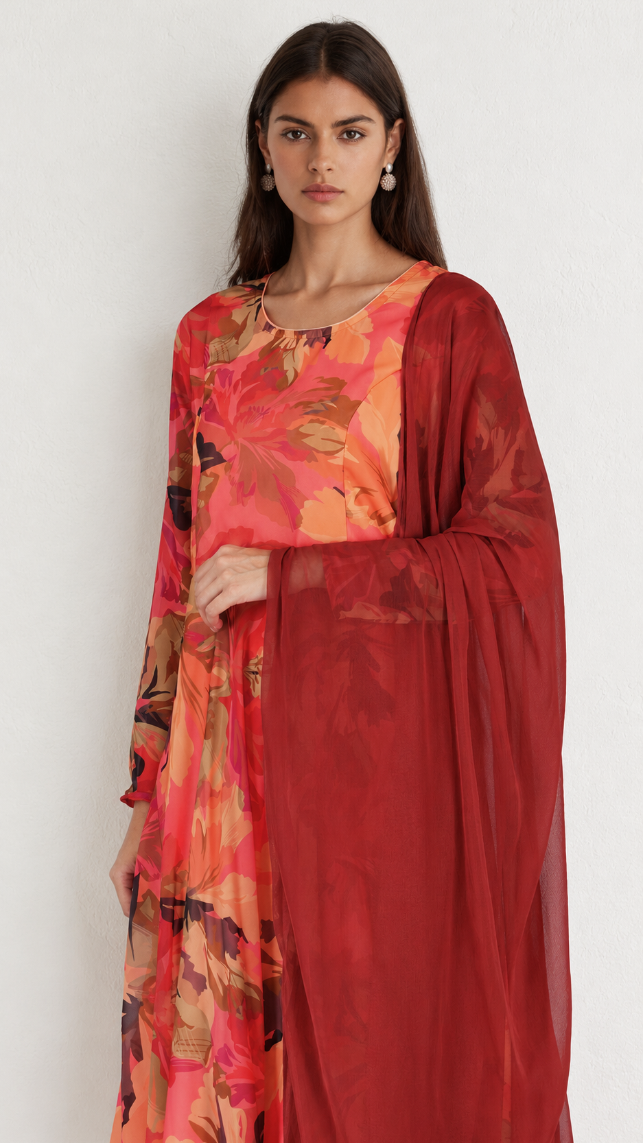 A woman modeling a traditional Indian outfit featuring a vibrant floral printed kurta in shades of orange, pink, and red, paired with a solid deep red sheer dupatta.
