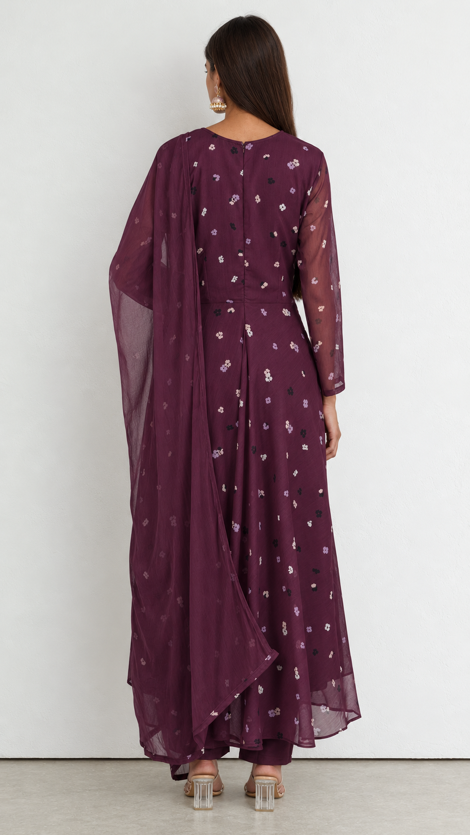 A woman viewed from the back wearing a deep purple anarkali-style dress with a matching dupatta and pants, decorated with small white floral prints, set against a plain light gray wall.
