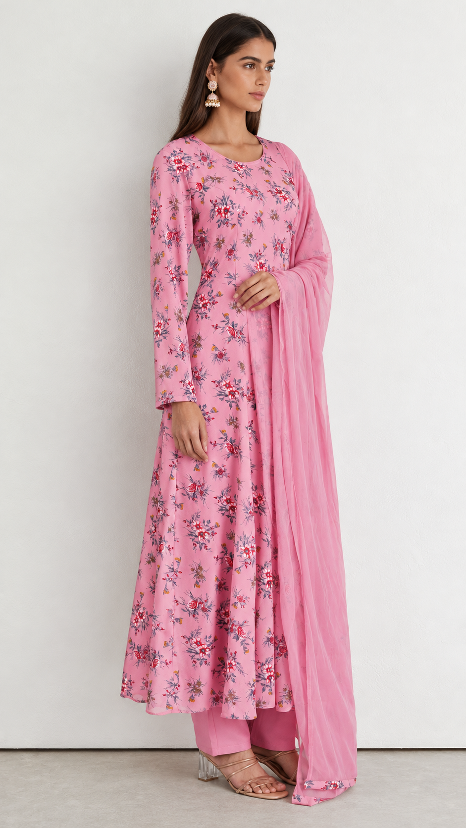 A woman modeling a pink long-sleeve kurta set with a floral print and a matching sheer dupatta, standing against a plain light gray wall.