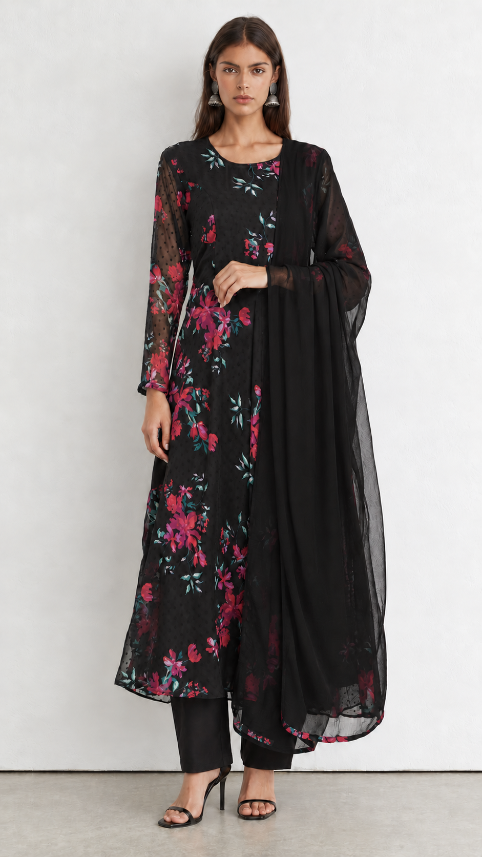 A woman modeling a black three-piece ethnic suit with a vibrant pink and red floral print, featuring a long kurta, matching pants, and a sheer black dupatta, set against a plain light gray studio background.