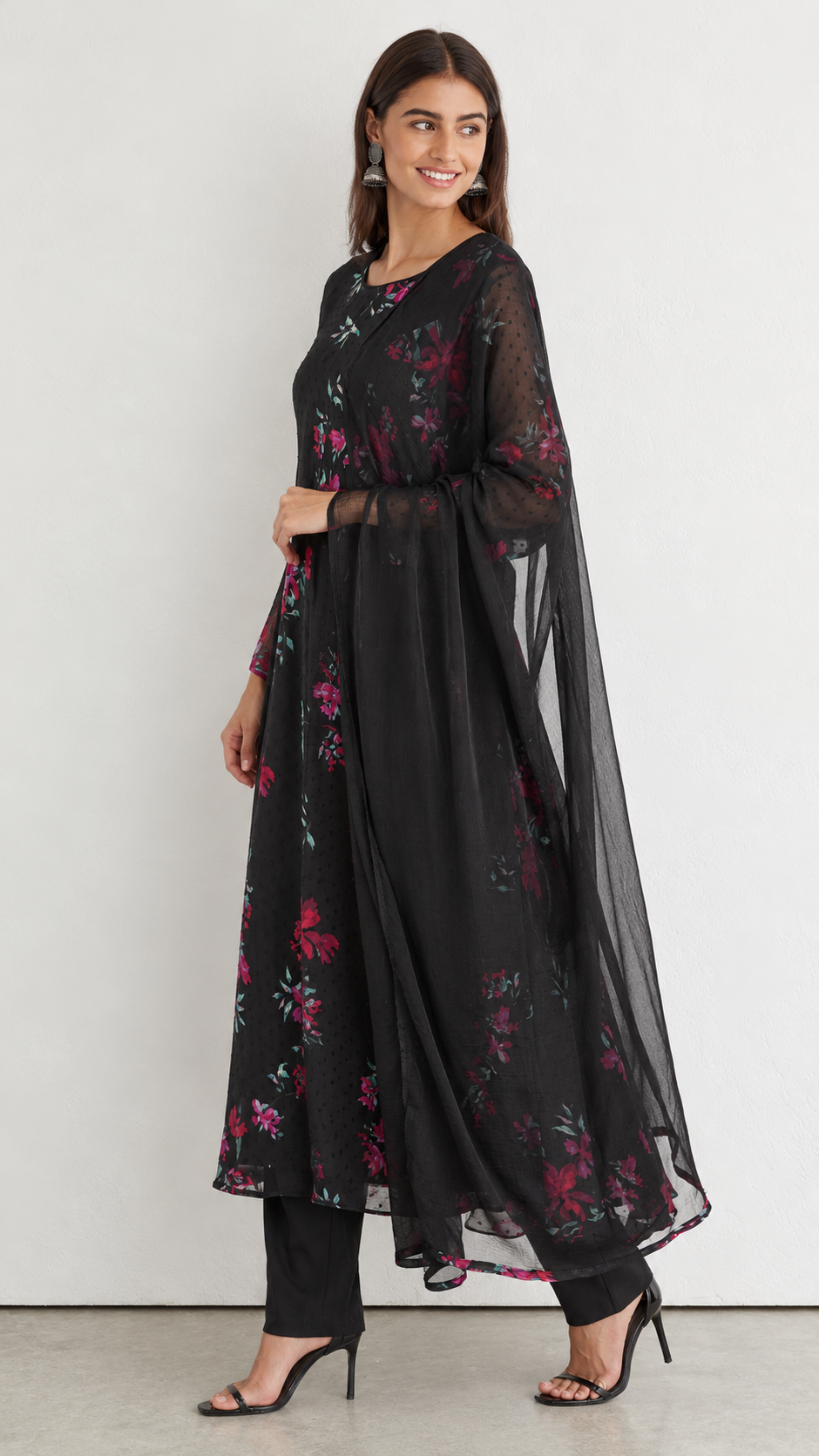 A woman models a black sheer floral kurta set with matching pants and a dupatta, featuring pink and red floral embroidery, against a plain light gray background.