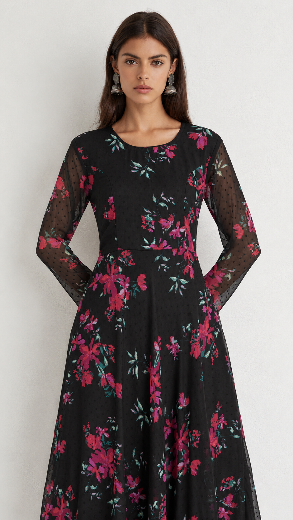 A woman modeling a black long-sleeve maxi dress with a vibrant pink and green floral print, featuring sheer mesh sleeves, set against a plain light gray background.