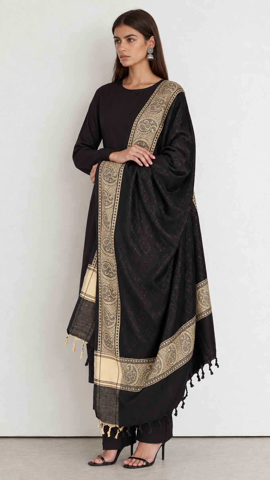 A woman modeling a black traditional outfit with a large black dupatta featuring wide beige and gold printed borders, standing against a plain light gray studio background.