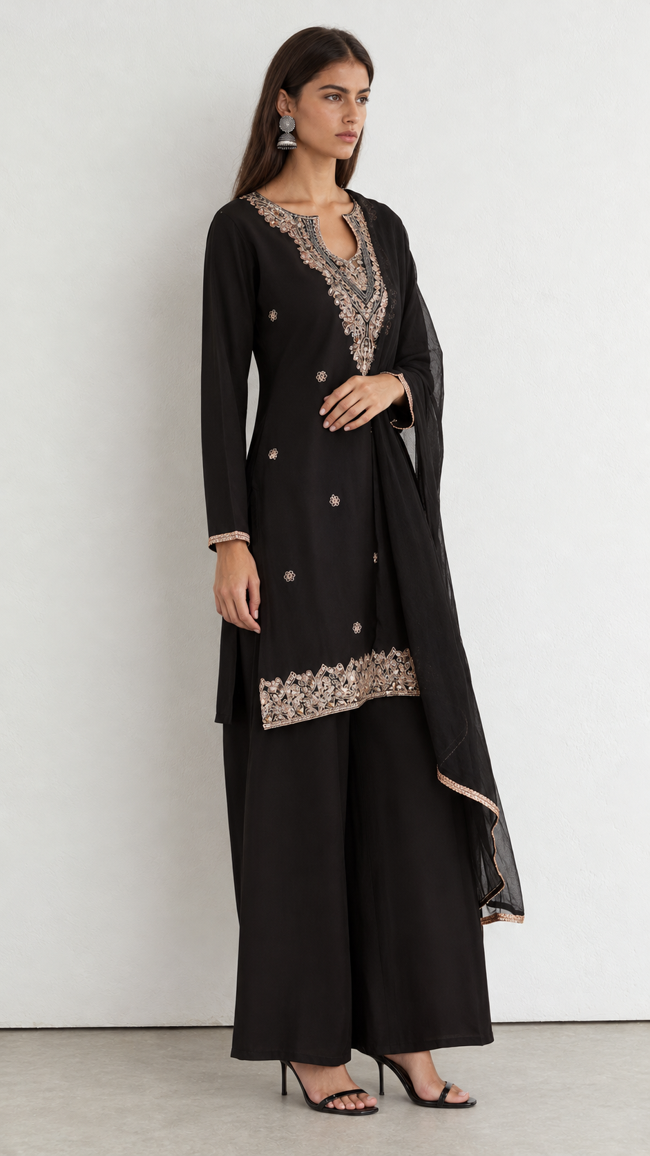 A woman modeling a black traditional outfit with beige floral embroidery on the neckline, hem, and dupatta, paired with wide-leg black pants and black heels, set against a plain light gray studio background.