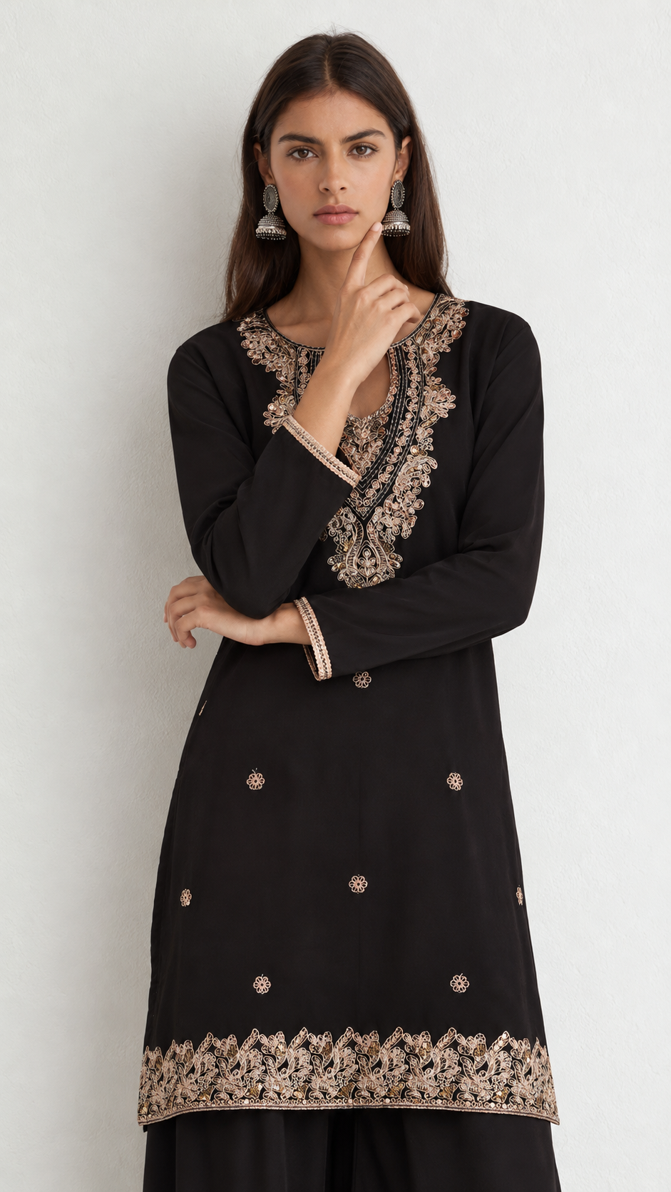 A woman modeling a black long-sleeve kurta with ornate beige and gold floral embroidery around the neckline, cuffs, and hem, paired with black pants against a light gray studio background.