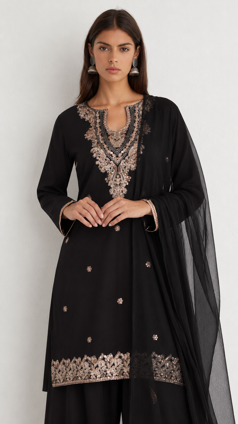 A woman modeling a black traditional outfit with ornate beige and metallic embroidery on the neckline, hem, and scattered decorative accents, paired with a sheer black dupatta against a plain light background.