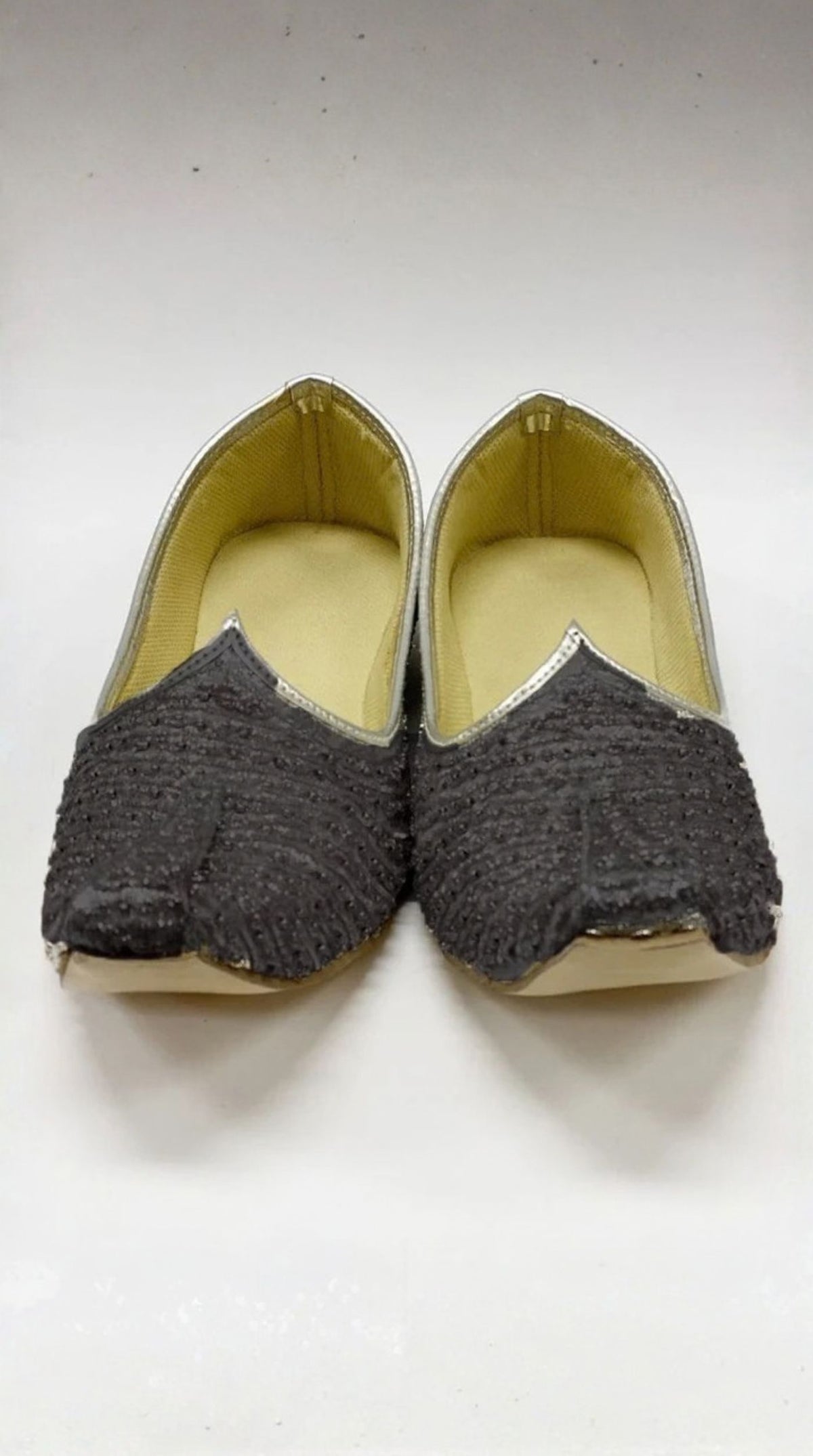 A pair of traditional Indian pointed-toe flat shoes, likely Jhumkas, featuring dark gray textured fabric uppers with silver metallic trim and a light beige insole.