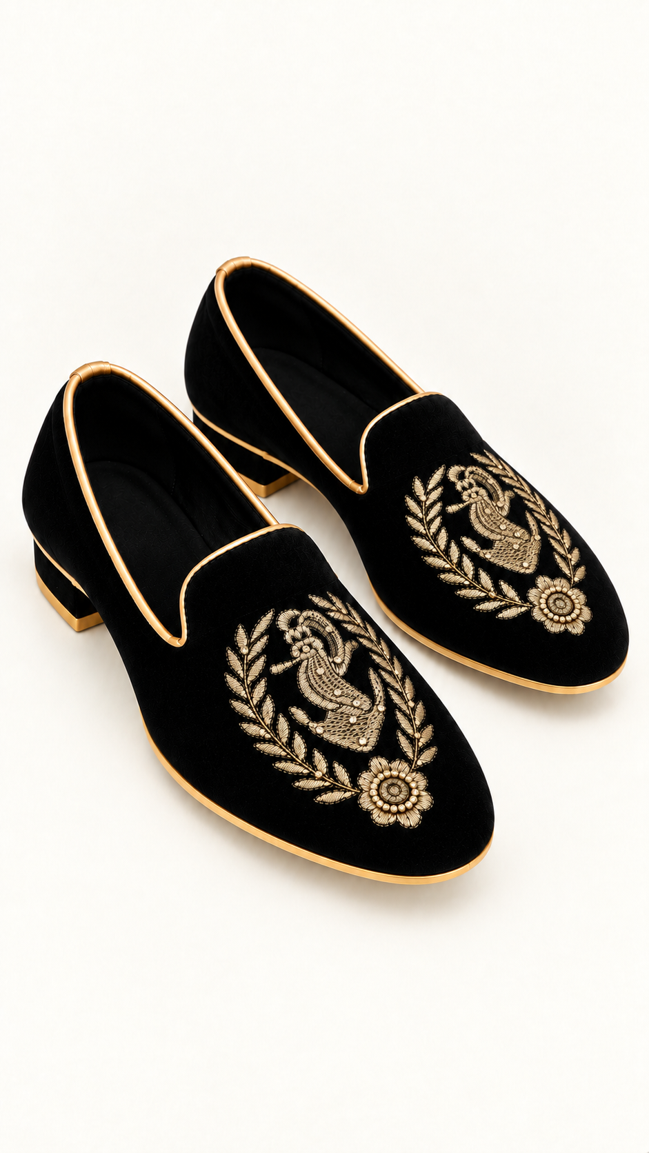 A pair of black velvet slip-on loafers with gold embroidered  laurel wreath motifs, accented with gold piping and low gold block heels, shown on a white background.