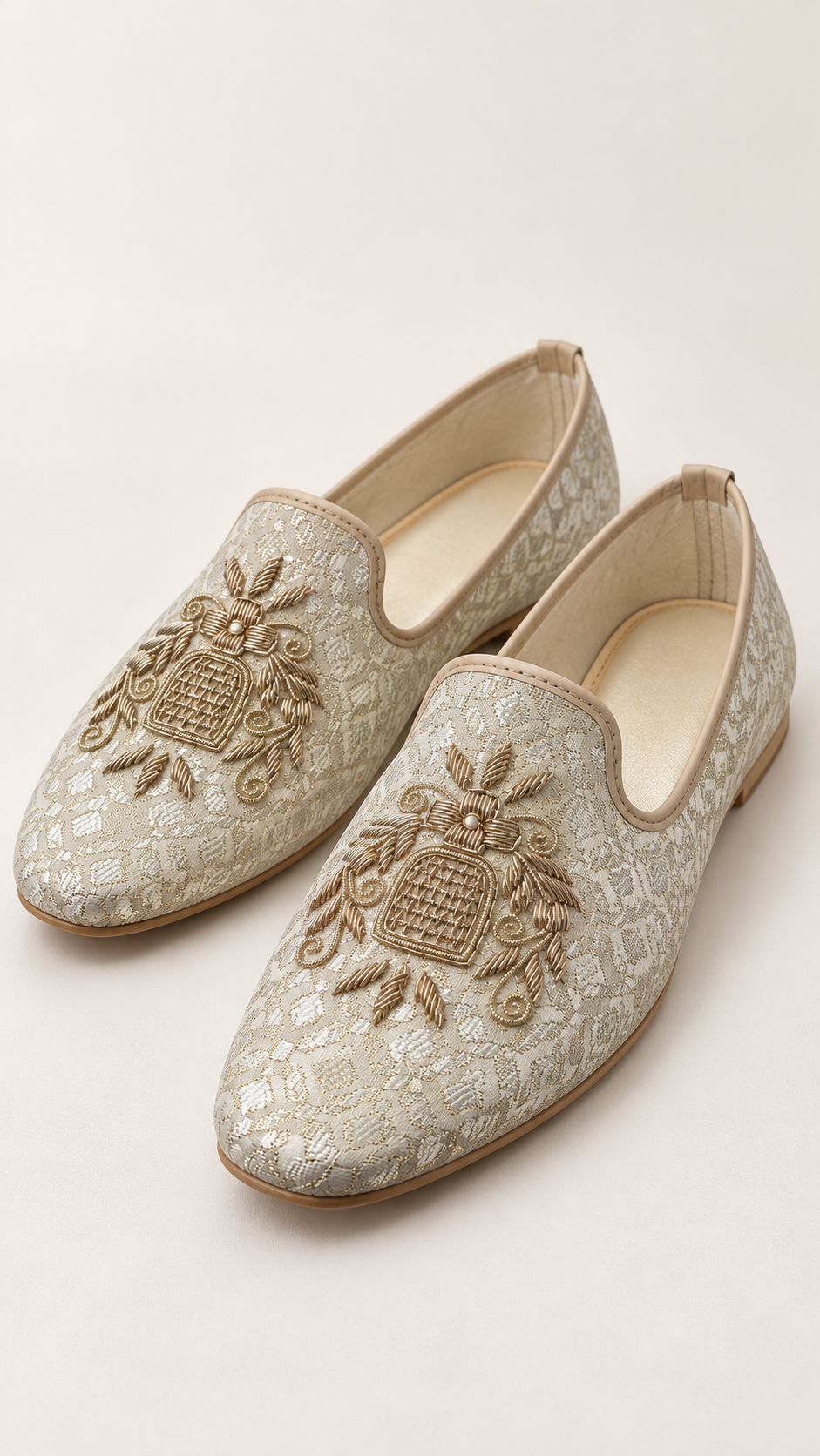 A pair of beige and gold embroidered slip-on loafers with a textured pattern and ornate decorative motifs on the toe, shown on a light beige background.