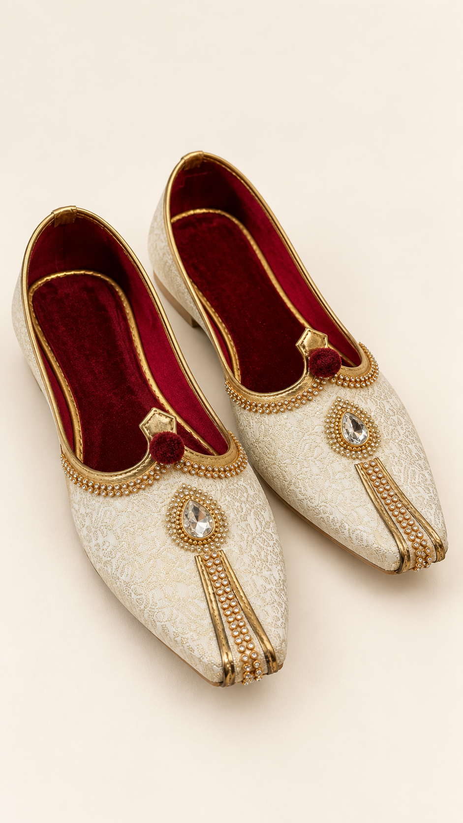A pair of traditional white embroidered sherwani shoes with gold trim, red velvet lining, and crystal embellishments, displayed on a light beige background.