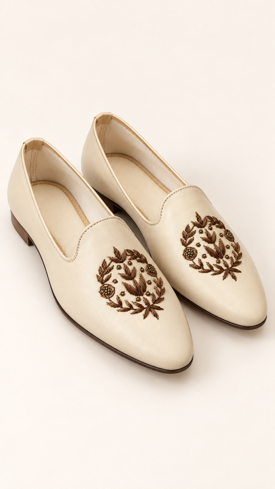 A pair of cream-colored leather slip-on loafers with brown embroidered floral wreath motifs on the toe, displayed on a light cream background.